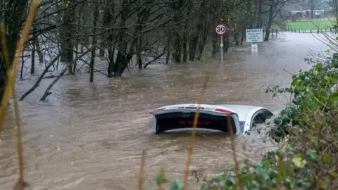 Great Impressions/Weather Watchers Grey car with its boot open nearly fully submerged in brown water on the road. There are trees on either side and a '30' sign and a sign that says 'Sedgwick'