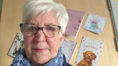 A woman with short white hair and black tortoiseshell glasses is taking a selfie.
There is a wooden door behind her with birthday cards stuck to it. She is wearing a blue and white scarf
