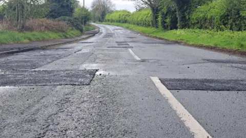 A low to the ground close up shot of a road, which is clearly in a damaged state and is covered in small patches where pothole repairs have taken place. The road is surrounded by hedgerows
