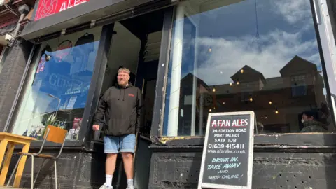 A man in a black hoody and blue denim shorts and trainers is standing in the doorway of a shop front which has two large glass windows either side of him and a black frame. There's an advertisement board at the front of the shop and a chair and wooden table in the left hand corner. The windows of the shop are reflecting buildings from the opposite side of the road.