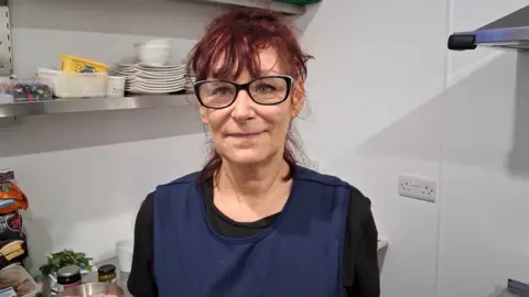 A women with red dyed hair, black glasses, smiling at the camera whilst wearing an apron stood inside a kitchen