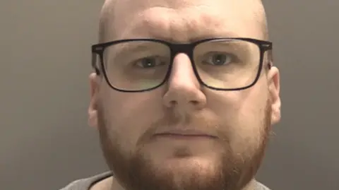 A police mugshot of Jack Mason, 31, who has a bald head, a ginger beard and is wearing black rimmed spectacles