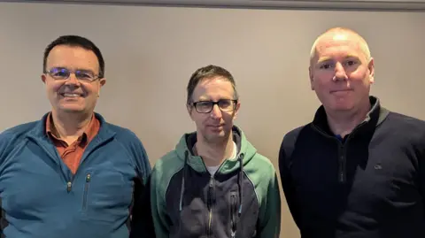 Handout Three men look at the camera, the first is wearing a blue sweater and orange t-shirt, the second is wearing a blue and green sweater, and the third is wearing a dark top.