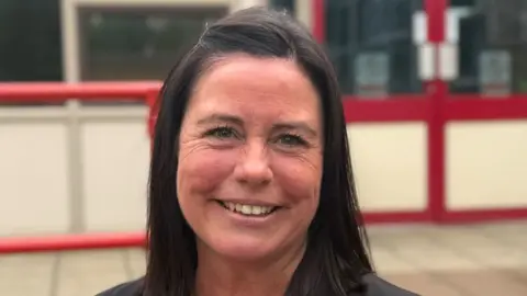 Elliot Deady/BBC A woman - Georgina Tatman - smiling straight at the camera. She is wearing smart clothes and standing in front of a school building.