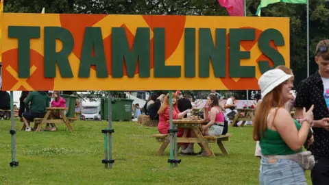 A large yellow sign in the grass reads "Tramlines' while young people sit at benches drinking next to it.