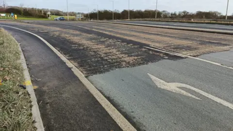 East Riding of Yorkshire Council A road with two lanes. The left lane had a white arrow pointed left painted on it. The road looks worn down. There is a grass verge on the left of the road and there is a roundabout in the distance.