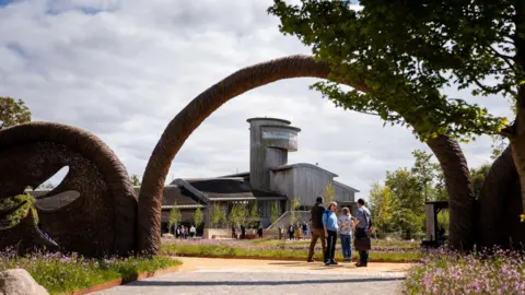 The Estuary Shoreline, a wetland landscape surrounded by wildflower meadows and native trees at Slimbridge. There is an arch which people are stood under. In the distance, a building can be seen, as well as lots of flowers and trees.