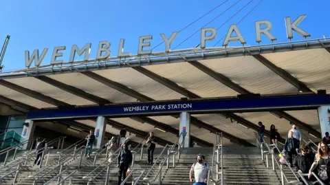 Marian McNamee/BBC The entrance to Wembley Park railway station. People are walking down a wide staircase that leads to the entrance.