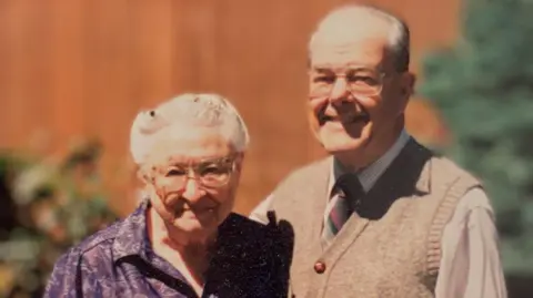 An old photograph of the late Dr Wally Baldwin and his wife, Wilma. They are smiling and there are trees in the background. 