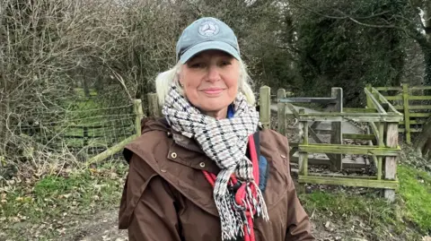 BBC/RICHARD EDWARDS Judy Noble - who remembers the disruption caused by the collapse of Tadcaster's bridge - is standing on the banks of the River Wharfe. A gate, leading to some woodland, is behind her. She is smiling at the camera and wearing a light-green baseball cap, a scarf and a dark jacket.