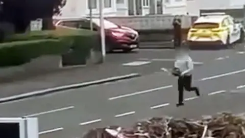 Grainy footage capturing a man wearing black trousers and a grey top running down a residential street while brandishing a chainsaw. A parked police car and a lone officer can be seen in the background