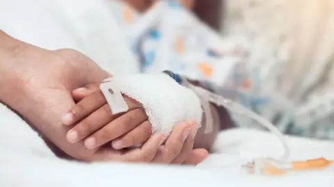 Getty Images A child's hand with a medical lead in, with a bandage, on a hospital bed. The child's hand is being held by an adult. 