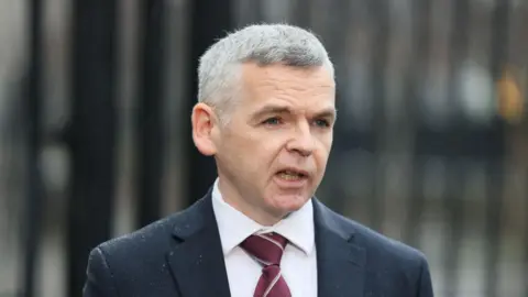 Pacemaker A man, with grey hair, wearing a navy suit, white shirt and burgundy tie. He is also wearing glasses and is holding a clip board and glasses. He is looking at the camera. 