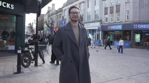 A man wearing a grey trench coat stands in the middle of a street. Various shops can be seen behind him, as well as bikes.