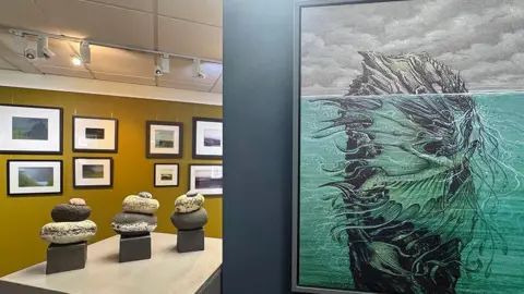 CAROLA RUSH An exhibition, featuring an illustration of a green dragon under water with its wings wrapped around a rocky piece of land that has a rocky top that is protruding above the water line. In the background there are other landscape paintings and clay sculptures.