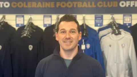 Stephen Fillis is a man with short dark hair.
His is standing in shop with hoodies on display behind him.
He is wearing a navy quarter zip with a lighter blue t-shirt underneath.