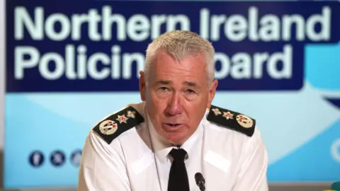 PA Media A grey haired man wears a white shirt, black tie with black patches on each shoulder. Behind him is a blue screen with the words "Northern Ireland policing board" on it.
