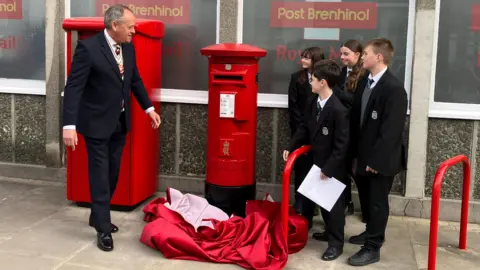 Four school children and a man with grey hair in a black suit stood in front of a red postbox carrying Kind Charles' insignia with a red cloth on the floor 