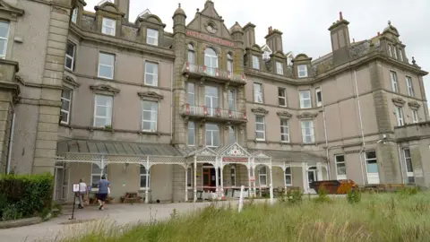 A multi-story building labeled "HOTEL VICTORIA" with numerous windows and chimneys. The entrance features a glass-covered area with white decorative elements and outdoor seating. Two people walk nearby, with grass visible in the foreground.