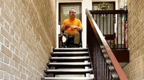 James Smith, a man in a wheelchair, is sat in the open front door of his flat at the top of a flight of stairs. The stairs are open stairs - so there is space between each one - and a wooden handrail is to the right. 