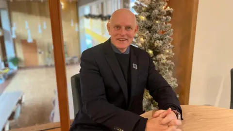 BBC / Gem O'Reilly Nick Phillips, who is wearing a black suit, is sat at a table in front of a Christmas tree and a window which overlooks an open indoor space. He is looking at the camera and smiling, with his hands clasped, resting on the table in front of him.