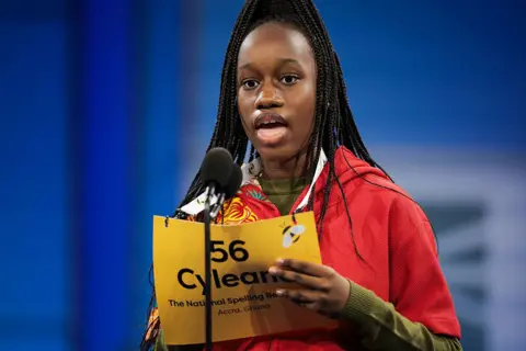 KAYLA BARTOWSKI / GETTY IMAGES A girl stands on stage and speaks into a microphone. She holds a yellow board showing her name, country of origin, and the National Belling Bee branding.