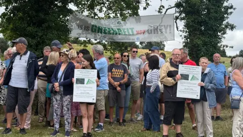 A gathering of people next to the land in Kingswinford known as The Triangle. Some people are holding signs opposing the development plans, with a banner in the background saying "this is green belt not grey".
