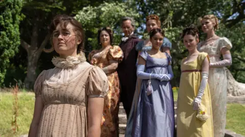 BBC | Bad Wolf | James Pardon The Bennet family, except Mary, stand on steps outside in a garden. Mary stands closer to the camera, in a beige dress with a mesh high neck