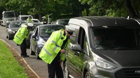 Multiple police officers in high-vis jackets stopping down cars stuck in traffic on a rural road asking to talk to them. 
