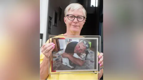 A woman wearing yellow holding a photo of her husband, who is cuddling a dog.
