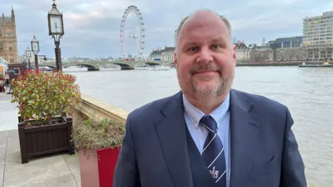 Jim Robbins is standing by the Thames in a blue suit. Behind him is the London Eye.  