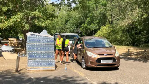 A distribution point for bottled water. Two women are seen next to a bronze car.