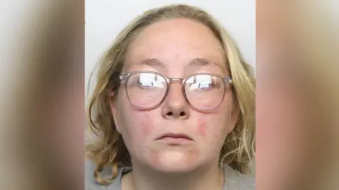 Greater Manchester Police Police custody image of Stacey Sharples who has shoulder length blond hair and pale pink rimmed glasses and wearing a grey t-shirt.