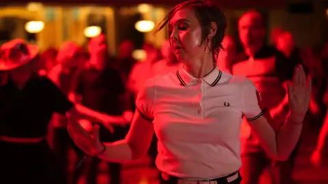 Getty Images A woman with short dark hair and wearing a white button-down T-shirt, dancing in the foreground with other dancers in the background at a Northern Soul event at Wigan Casino Club.