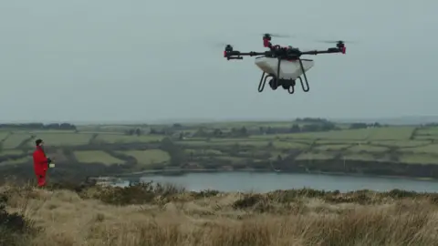 A landscape of a grey British moorland. A man in red overalls stands on the edge of a cliff. He is looking up at the sky towards a large drone. The drone is in the foreground of the image and has a white body and four black rotors. 