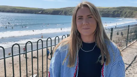 BBC Mirain Llŷn Roberts stands near the seafront in Aberdaron and behind her are people on the beach and in the water