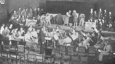 Bedales Record An image of the 1931 performance in the Holywell Music Room. In the foreground, older children are playing musical instruments. Up on stage, younger (upper primary school age) children are singing and acting. Some of them are holding props – including pieces of paper painted with bricks on, which is quite fun. It is a black and white image.