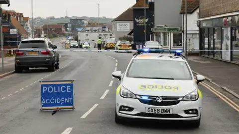 Eddie Mitchell A police car at the scene of the crash on a road that has been taped off with an officer standing guard