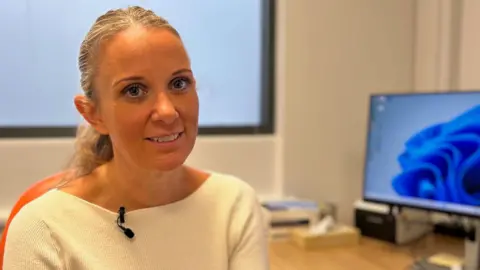 Alex has long blonde hair tied behind her head in a ponytail. She is wearing a plain while jumper and is pictured at her desk in one of her GP surgeries. 