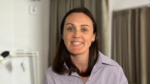 Steph is seated on the edge of a bed in a tidy bedroom, facing the camera, wearing a long-sleeved striped shirt. Behind her are closed floor-length curtains and a wall-mounted reading lamp positioned above a bedside table. The room has neutral walls, soft indoor lighting, and a calm, uncluttered appearance.