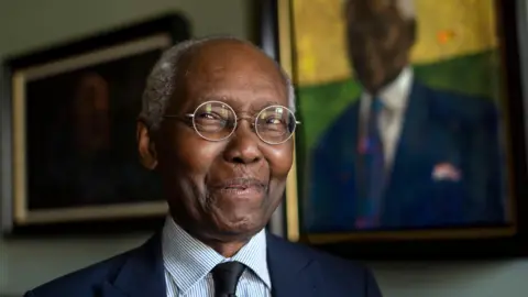 PA Media Sir Geoff Palmer, wearing a suit, glasses and with white hair, stands in front of a portrait of himself.