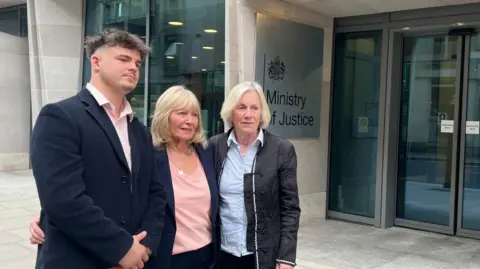 A man and two women stand shoulder to shoulder outside the Ministry of Justice building. All three are wearing smart clothing.