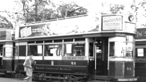 Brighton 53 Tram Society A black and white picture of tram 53 at the old steine in Brighton in 1938. It has advertising on the side and a sign that says "palace pier" on the side and the number 53 on the side with the company name "Brighton corporation tramways". A person is walking beside it
