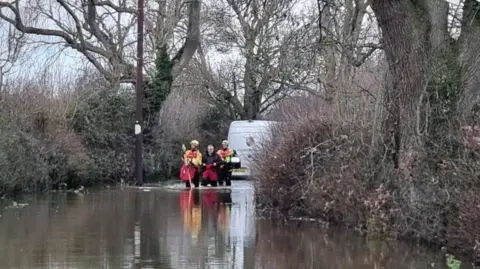 Shropshire Fire and Rescue Service Three men in red and black waterproof clothing standing in a flooded road with trees on either side and a white van behind that