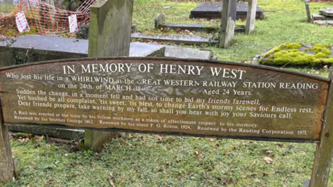 A wooden memorial stands in a graveyard and bears the inscription 'in memory of Henry West'