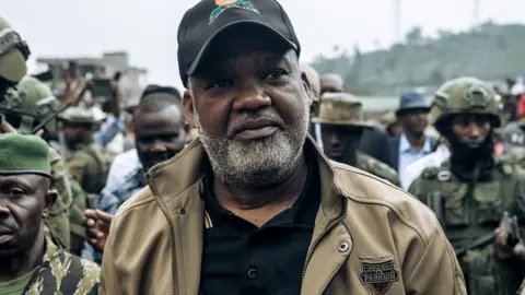 AFP Congo River Alliance Corneille Nangaa, wearing black cap, black T-shirt and brown jackets, arrives at the Stade de l'Unite in Goma on February 6, 2025. He is surrounded by M23 fighters.