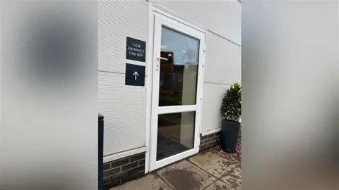 Steph Birch An exterior door that is white with two large glass panes. The door handle is three quarters of the way up the door. A sign next to the door says "club entrance this way" and another sign below it has an arrow pointing in the direction of the door