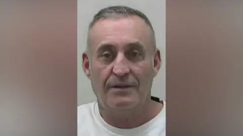 Mugshot of David Kelly. He is clean shaven with very short grey hair, cropped at the sides. He has a piercing in his left eyebrow and appears to have a small tattoo next to his left eye.