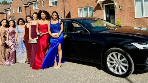 Women in long evening dresses line up alongside a long, black, shiny limousine parked outside a neat read brick house.
