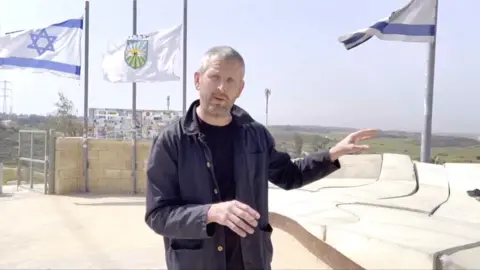 A man, Jon Donnison, at Sderot in Israel, with Israeli flags behind him and Gaza in the distance.